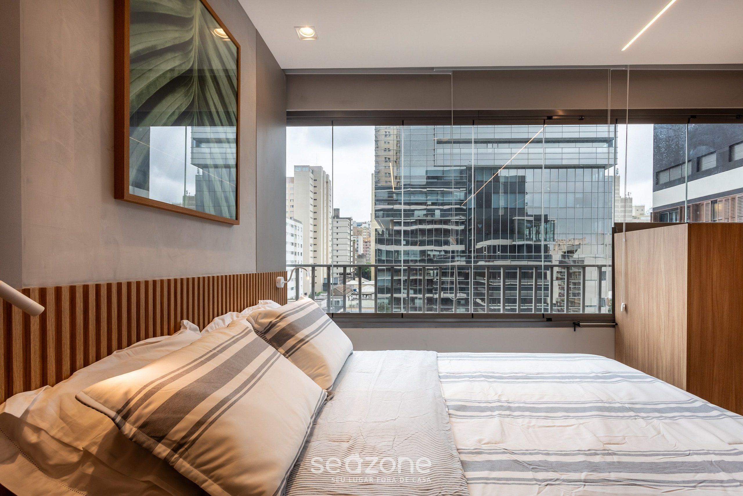 A modern bedroom with a large window, a bed with striped bedding, and a city view.