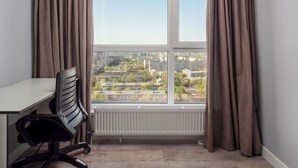 Et værelse med udsigt til bybilledet gennem et stort vindue, en radiator under vinduet, et skrivebord med en stol, og brune gardiner.