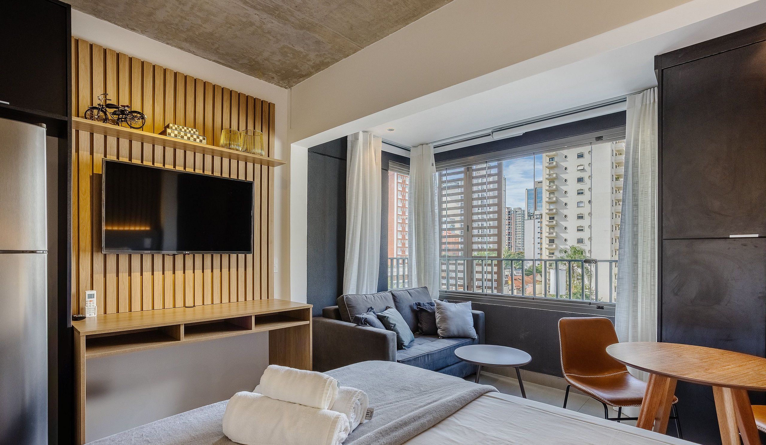 A modern hotel room with a flat-screen TV, a sofa, a small table, and a balcony with a city view.