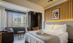 Ein modernes Hotelzimmer mit einem großen Bett, einem kleinen runden Tisch, einer Couch und Blick auf die Stadt durch große Fenster.
