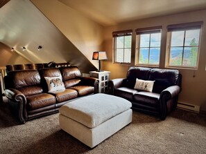 A cozy living room with a brown leather sofa, a light-colored ottoman, and a small table with a lamp. There are two windows with views of the outdoors. - New to VRBO! Ski-in/out in 15 Steps - Alpine Meadows Chalet - Hot Tub -Mtn Views (Big Sky)