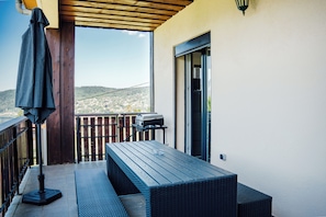 A balcony with a wooden table and bench, a grill, and a view of a town and hills. - Le Grand Duc: Lake view and close to downtown (Gérardmer)