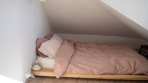 Ein Einzelbett in einem Loftsessel-Zimmer mit geneigter Decke, ein Nachttisch mit Lampe und ein wandmontiertes Regal.