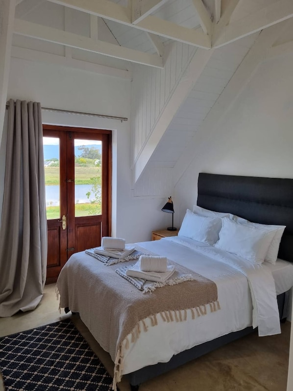 A bedroom with a bed, a nightstand, a lamp, and a view of the outdoors through a door.
