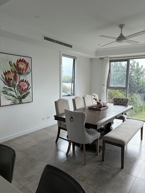 A dining area with a large table, six chairs, and a bench. There are two windows providing natural light and a view of greenery.