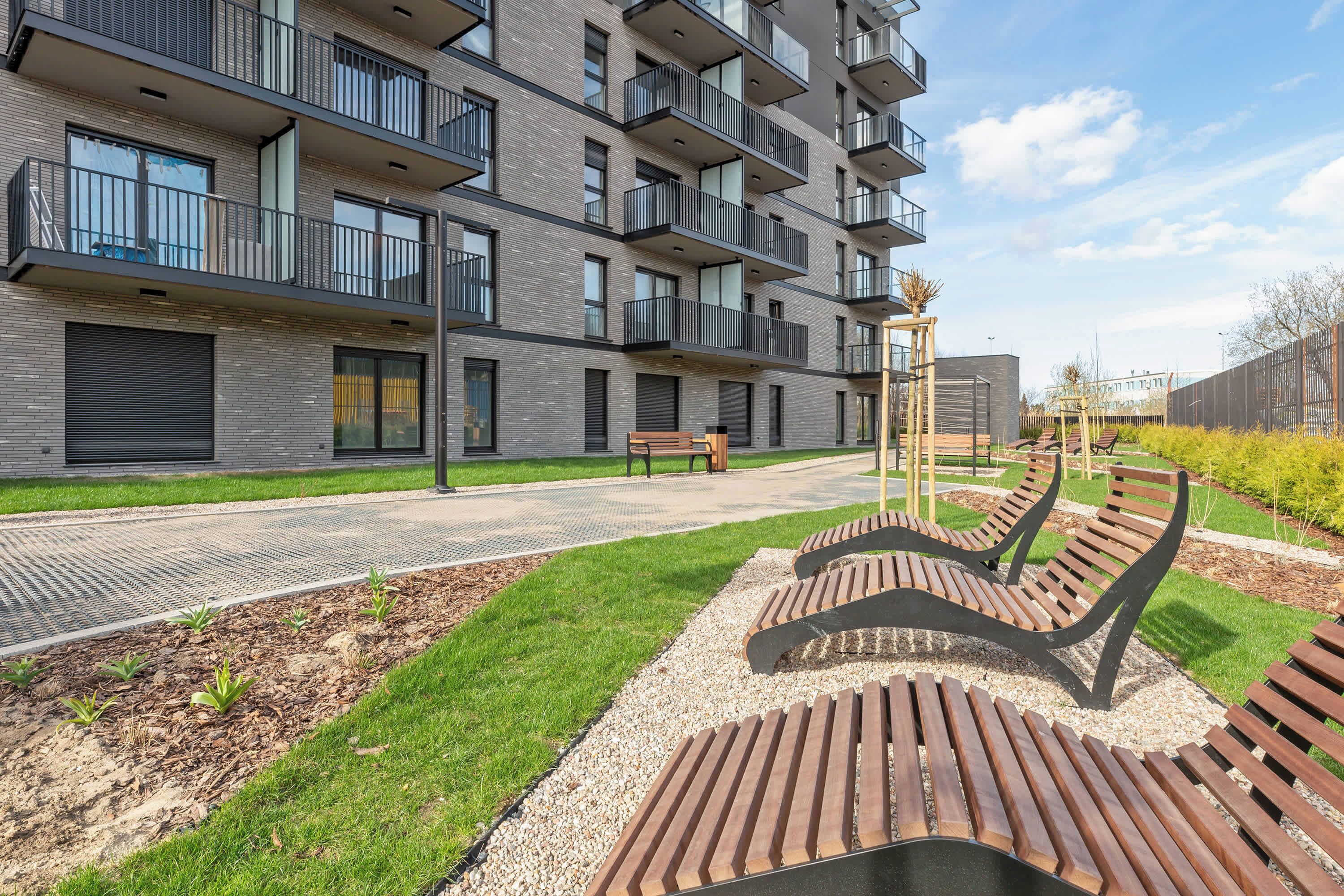 A modern multi-story residential building with balconies, a paved walkway, and a landscaped garden area with benches.