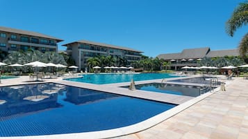 Uma grande piscina ao ar livre com várias pistas de natação, rodeada por espreguiçadeiras e guarda-sóis, e adjacente a um edifício de hotel com vários andares.