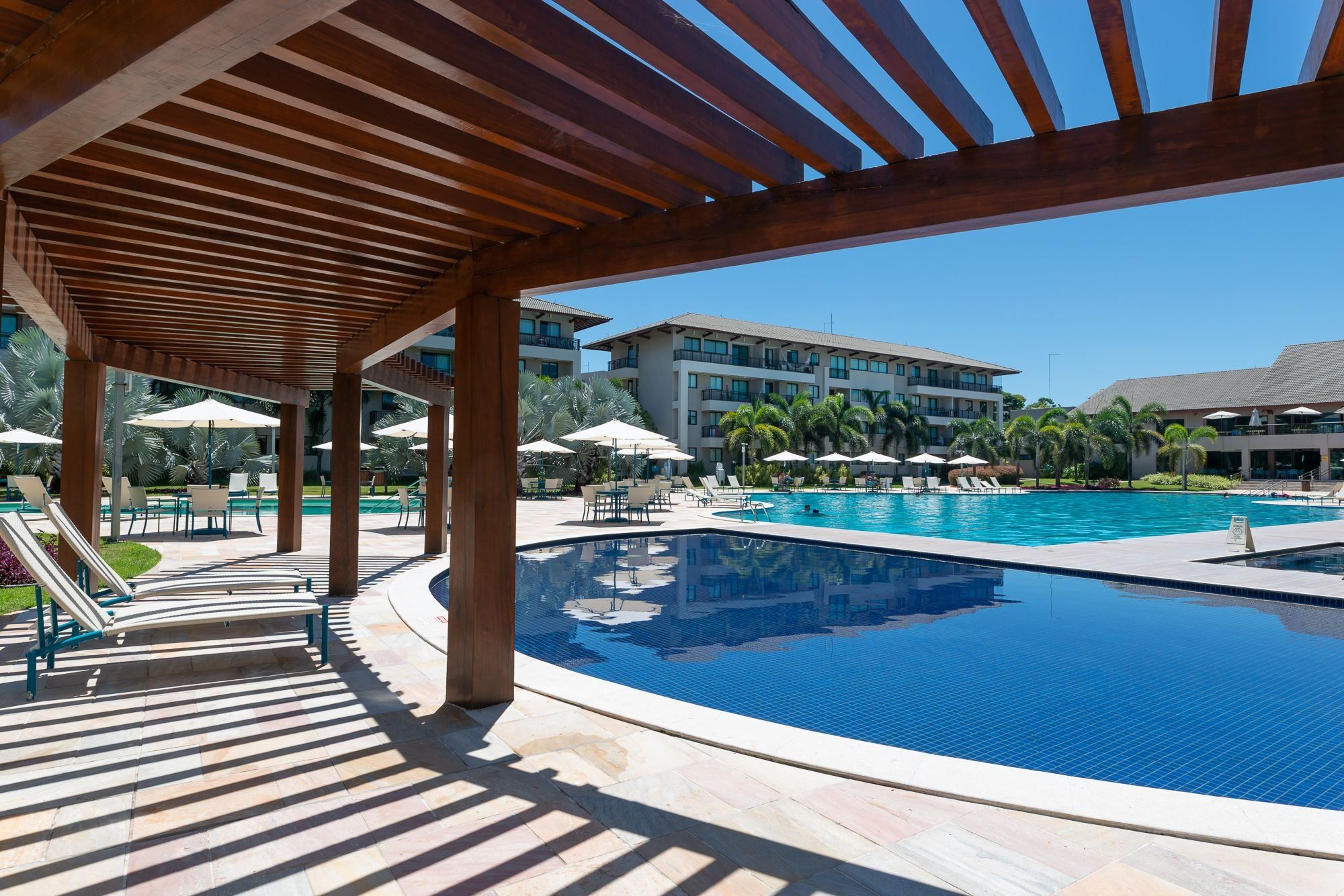 Área de piscina com pérgola de madeira, cadeiras de descanso e um edifício ao fundo.