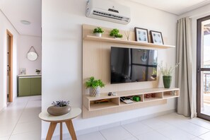 A modern living room with a flat-screen TV mounted on a wooden wall unit, a small round table with a plant, and a sliding glass door to the right. - BeachClass EcoLife Porto de Galinhas (Ipojuca)
