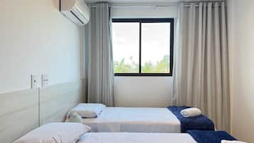 A hotel room with two beds, a window with curtains, an air conditioning unit, and a view of greenery outside.