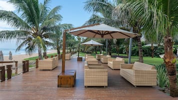 Terraza de madera con mobiliario de mimbre y sombrillas, con vista a la playa.