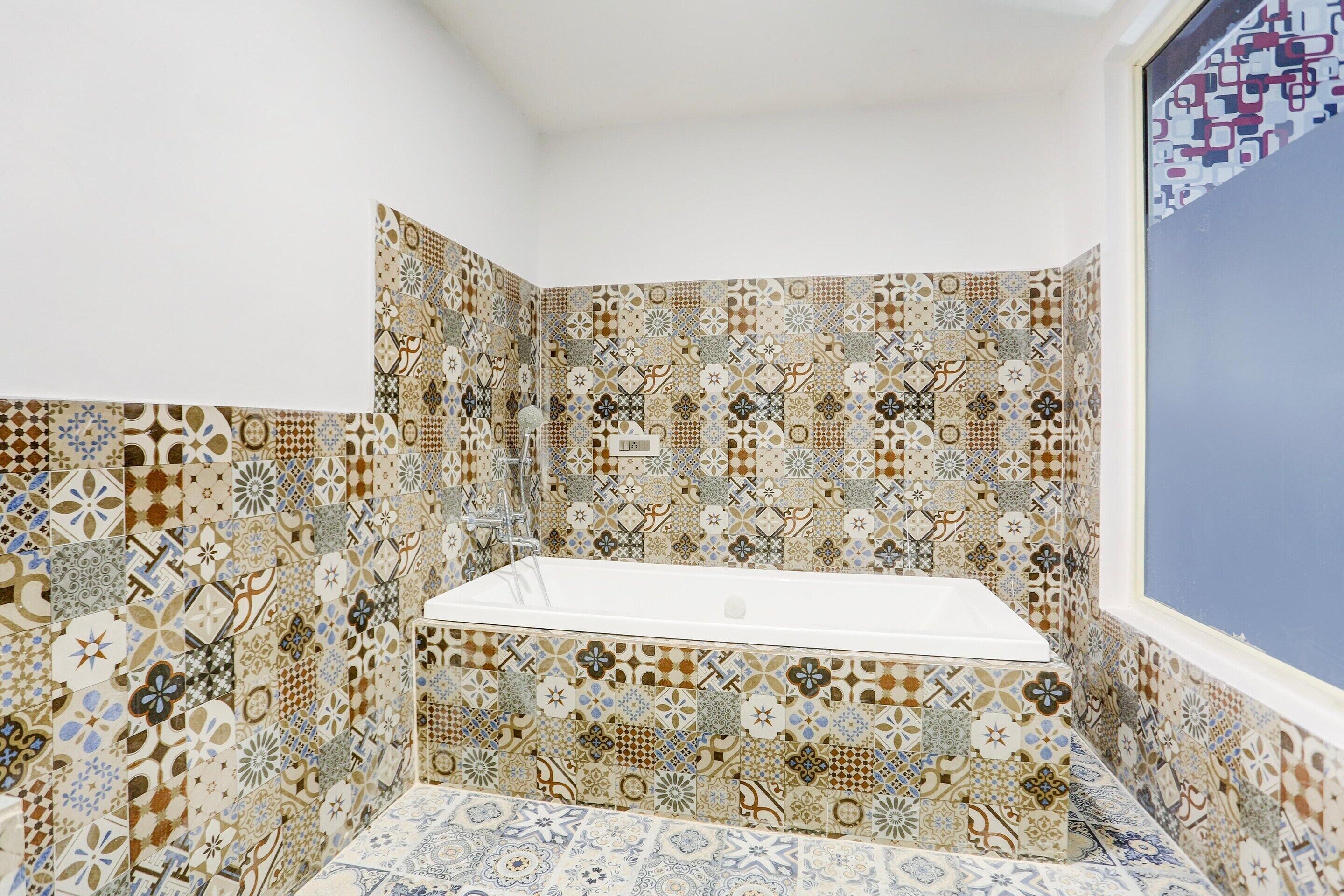 Casa de banho no canto com banheira branca, azulejos estampados e janela.