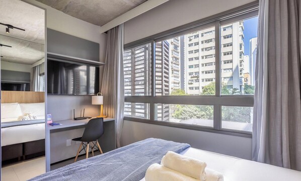 Ein Hotelzimmer mit Bett, Schreibtisch, Stuhl, Lampe und einem großen Fenster mit Blick auf hohe Gebäude.