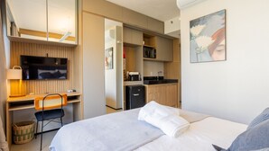 A hotel room with a bed, a desk with a chair, a television, a kitchenette, and a painting on the wall. - Beautiful Studio 800m From the Metro Oag0505 (São Paulo)