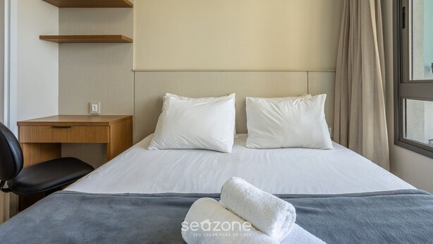 Ein Hotelzimmer mit Bett, zwei Kissen, einem Nachttisch, einem Sessel und einem Fenster mit Vorhängen.