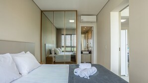 Ein modernes Hotelzimmer mit einem großen Bett, einem begehbaren Kleiderschrank, einem Badezimmer mit Waschbecken und Spiegel sowie Blick ins Freie.