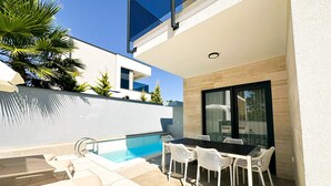 Uma área de piscina moderna ao ar livre, com uma mesa e cadeiras de jantar, cercada por uma parede branca e uma porta de vidro.