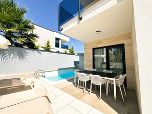 Ein moderner Poolbereich im Freien mit Esstisch und Stühlen, umgeben von einer weißen Mauer und einer Glastür.