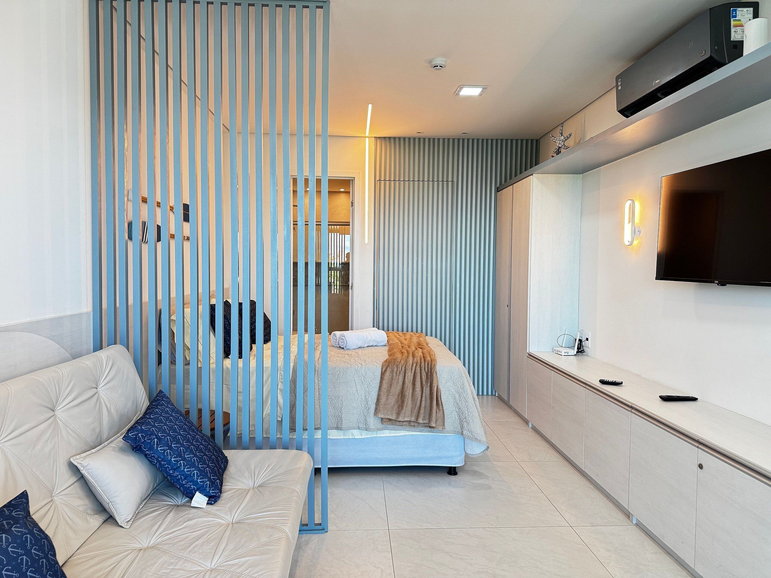 A modern hotel room with a bed, a sofa, a TV, and a partition with vertical slats.