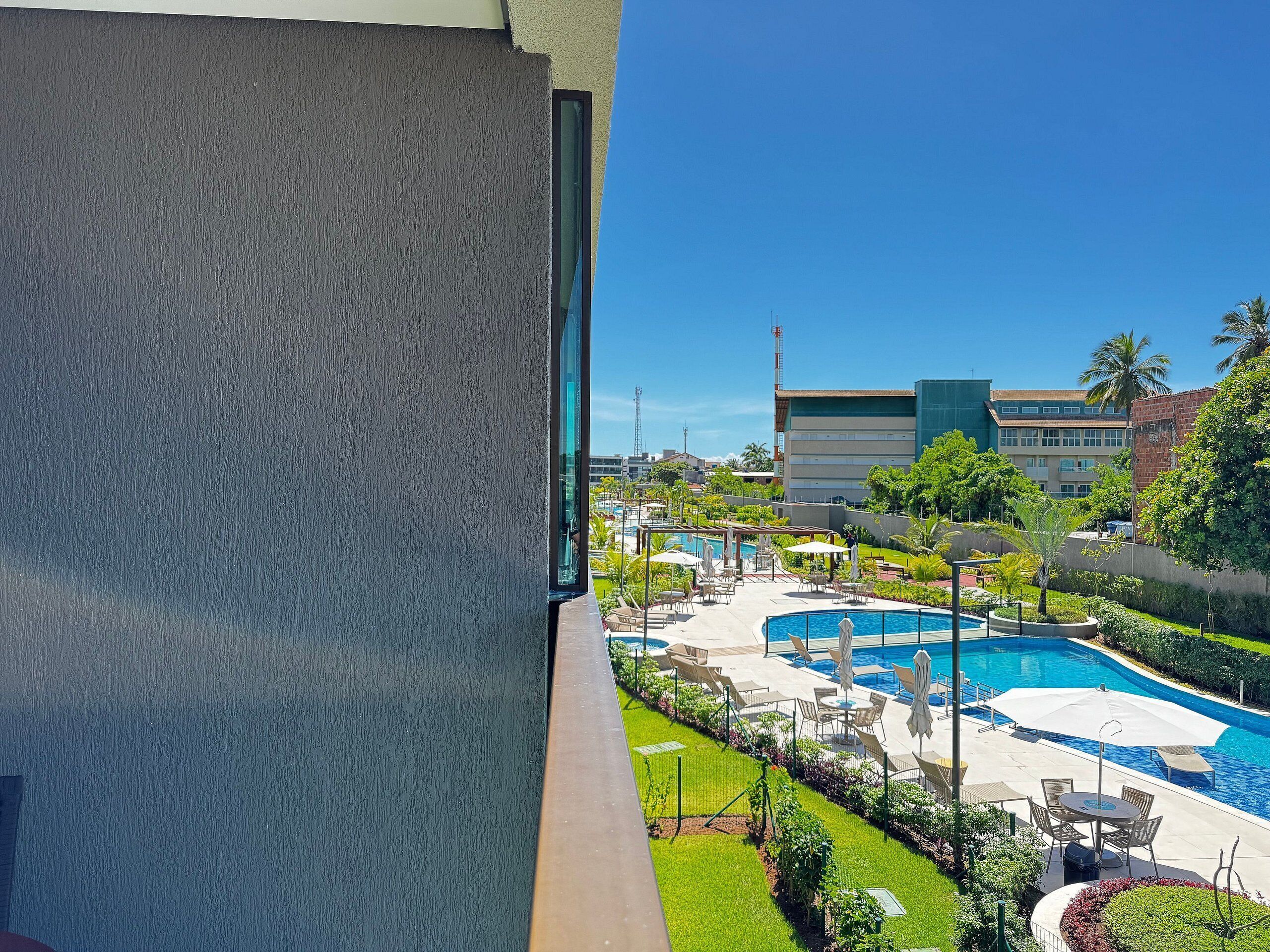 Uma área de piscina com espreguiçadeiras e guarda-sóis, cercada por vegetação e edifícios ao fundo.