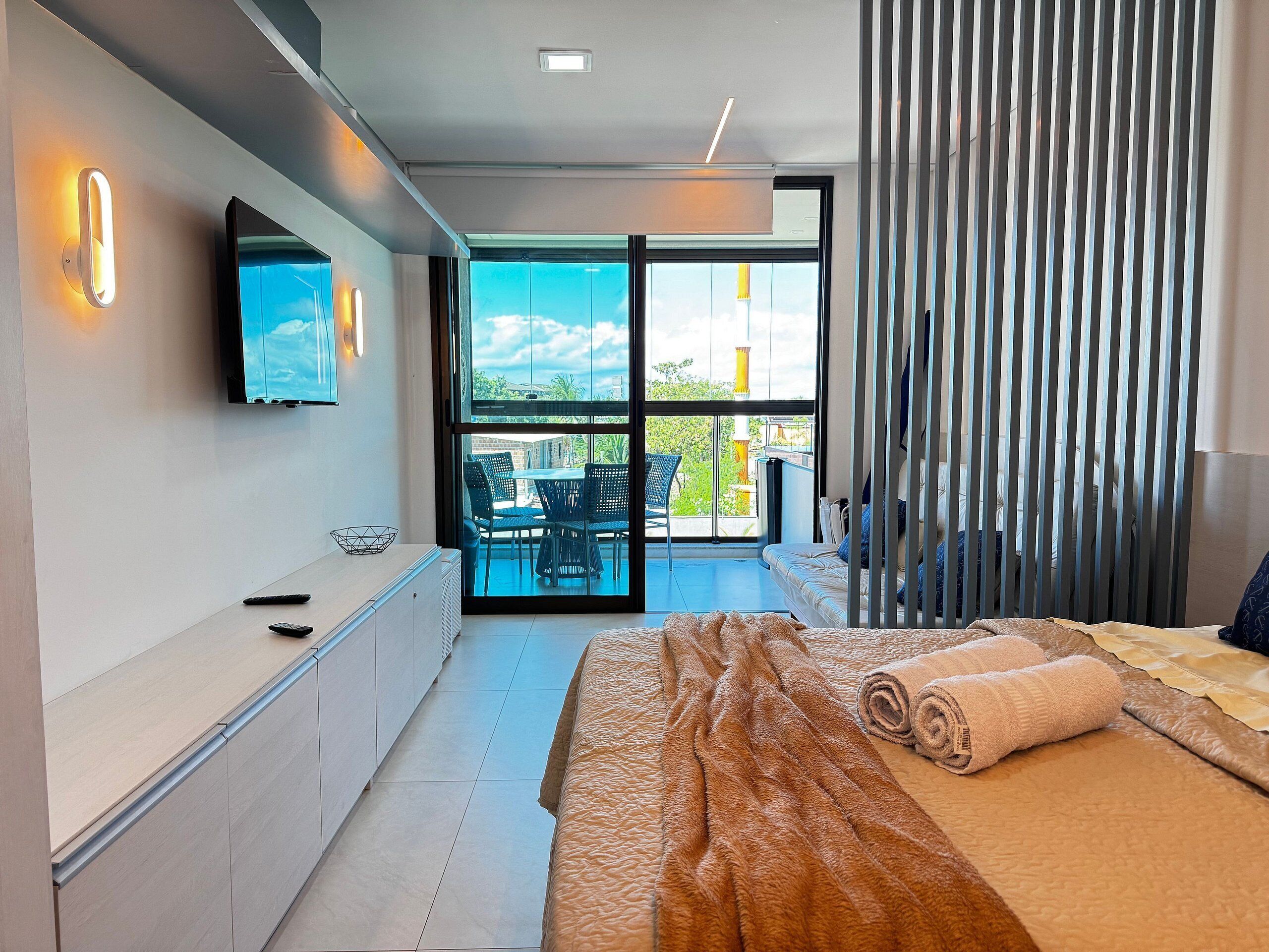 Un dormitorio moderno con una cama grande, un televisor montado en la pared y un balcón con mobiliario exterior.