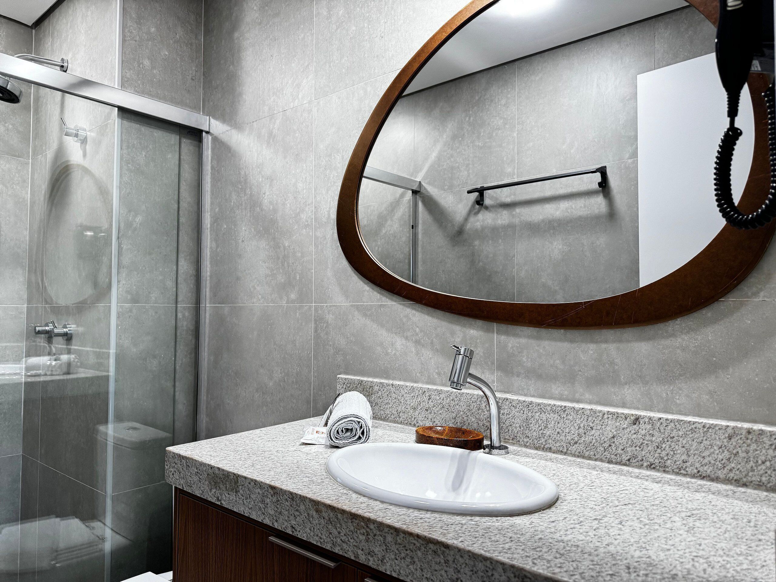 Un baño con cabina de ducha de vidrio, un espejo redondo con marco de madera, un lavamanos blanco con un jabonera de madera y un dispensador de toallas.