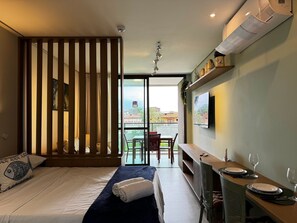 Habitación de hotel con cama, zona de comedor con mesa y sillas, vista del paisaje urbano y televisión montada.