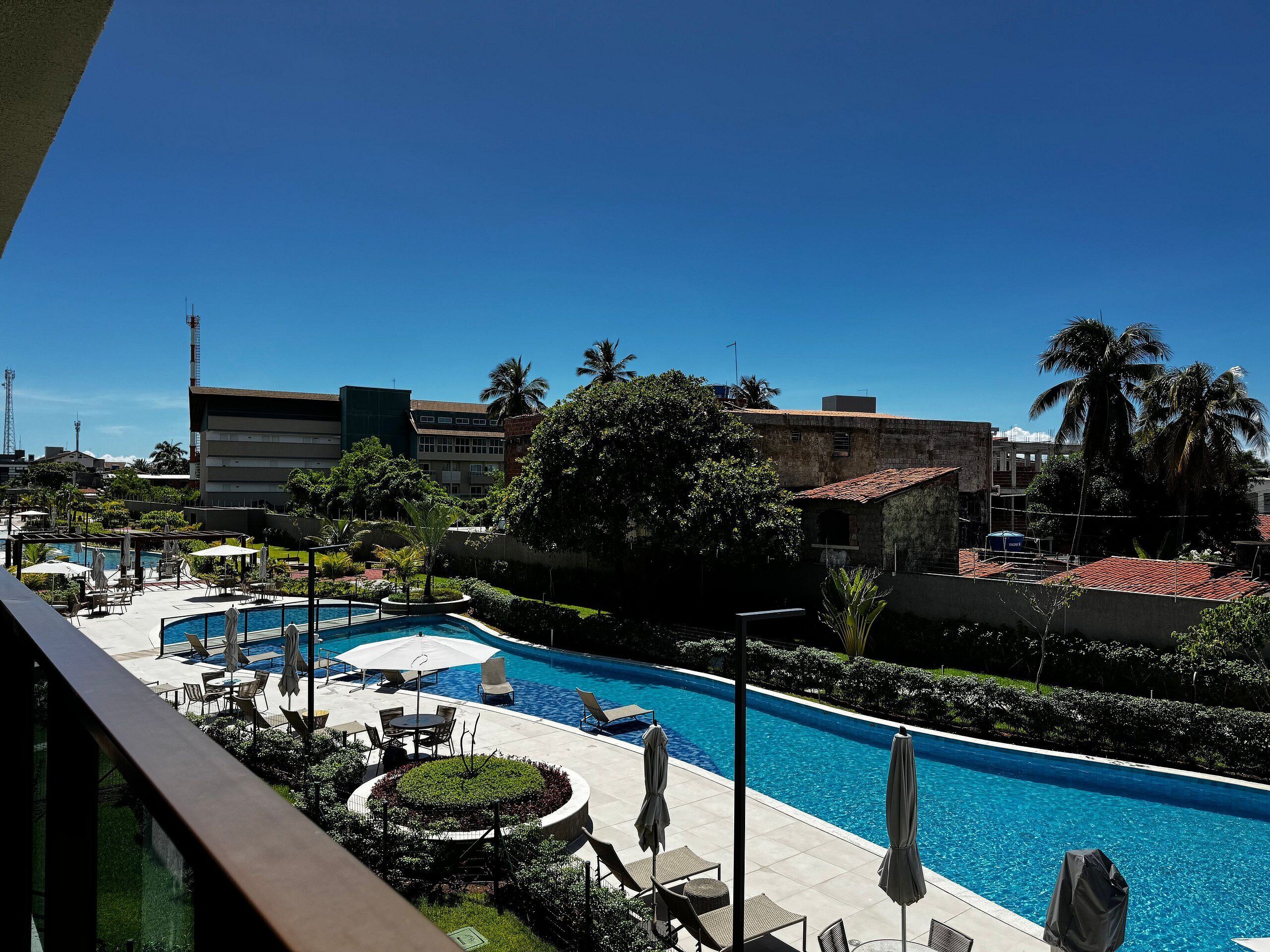 A hotel pool area with lounge chairs and umbrellas, surrounded by greenery and buildings.