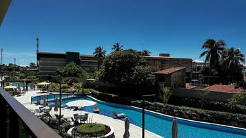 A hotel pool area with lounge chairs and umbrellas, surrounded by greenery and buildings.