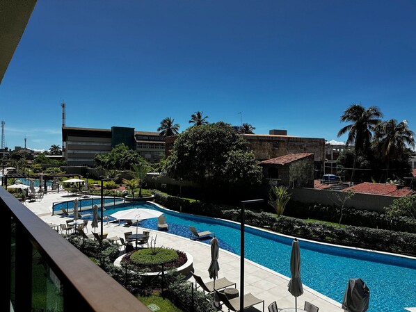 Zona de piscina hotelera con sillones de descanso y sombrillas, rodeada de vegetación y edificios.