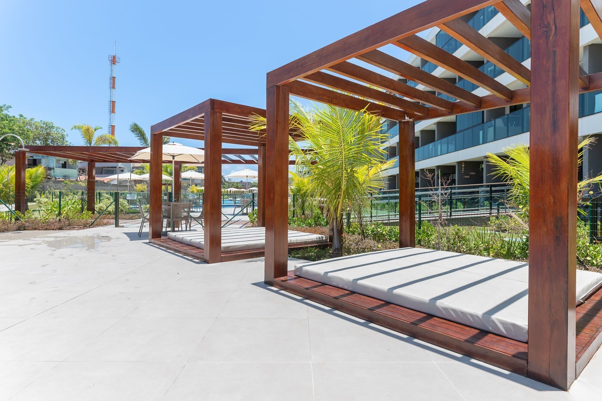 Un espacio de estar al aire libre techado con pérgolas de madera, una piscina y un edificio moderno al fondo.