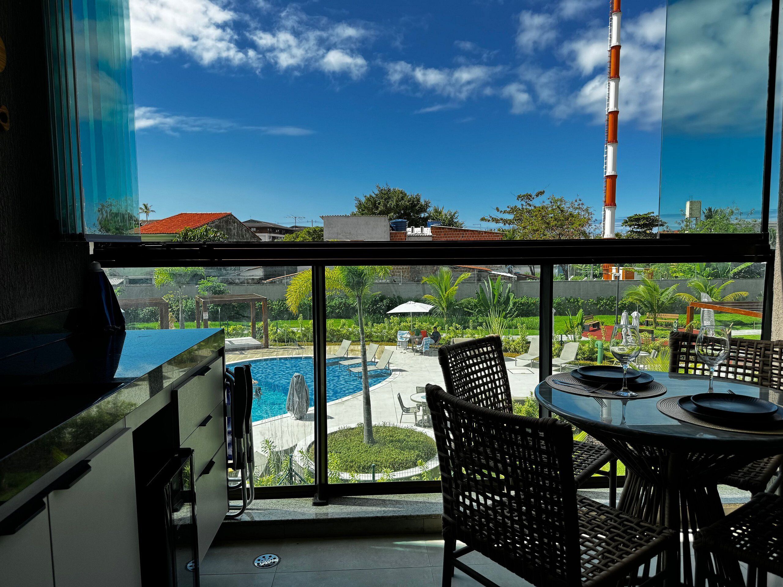 A balcony with a view of a swimming pool, outdoor seating, and a clear sky.