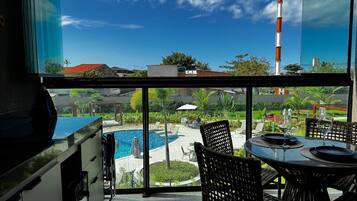A balcony with a view of a swimming pool, outdoor seating, and a clear sky.
