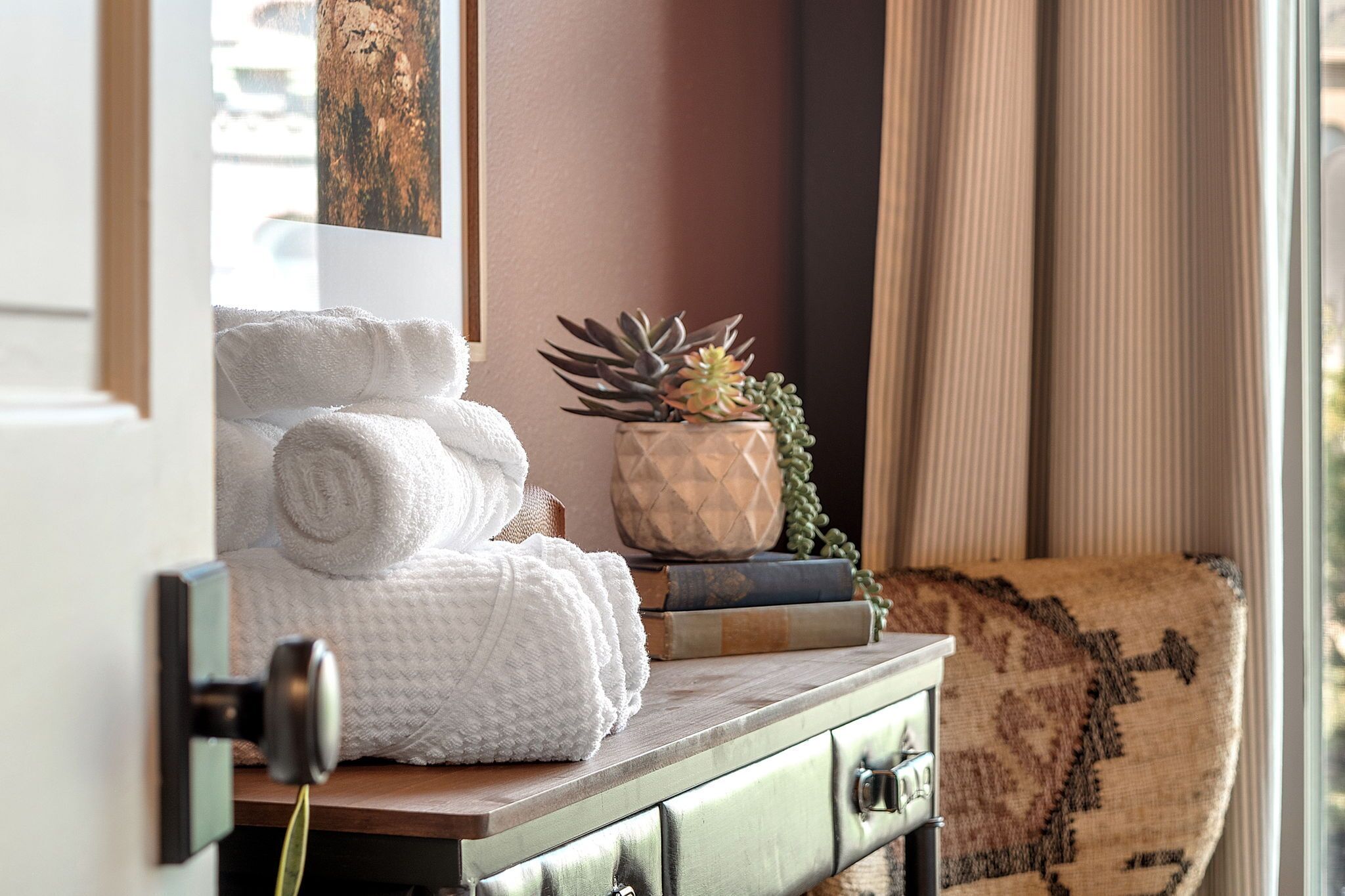 A room with a wooden dresser, a stack of white towels, a potted succulent, and a patterned throw on a couch.