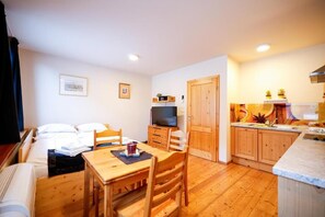 A compact room with a bed, a small dining table with chairs, a television, and a kitchenette with wooden cabinets.