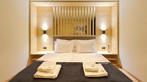 Quarto de hotel com cabeceira de madeira, duas mesas de cabeceira, cama com roupa de cama branca e duas luminárias fixadas na parede.