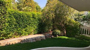Un espacio de descanso al aire libre con asientos curvos y una superficie reflectante, rodeado por un jardín cuidado con diversas plantas y un muro de piedra.