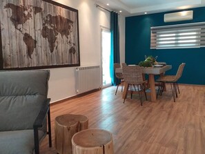 A modern living room with a sofa, a dining table with chairs, a world map artwork, a radiator, and a window with blinds. - Casa Panorama in Voula - Panoramic Sea View Villa (Vari-Voula-Vouliagmeni)