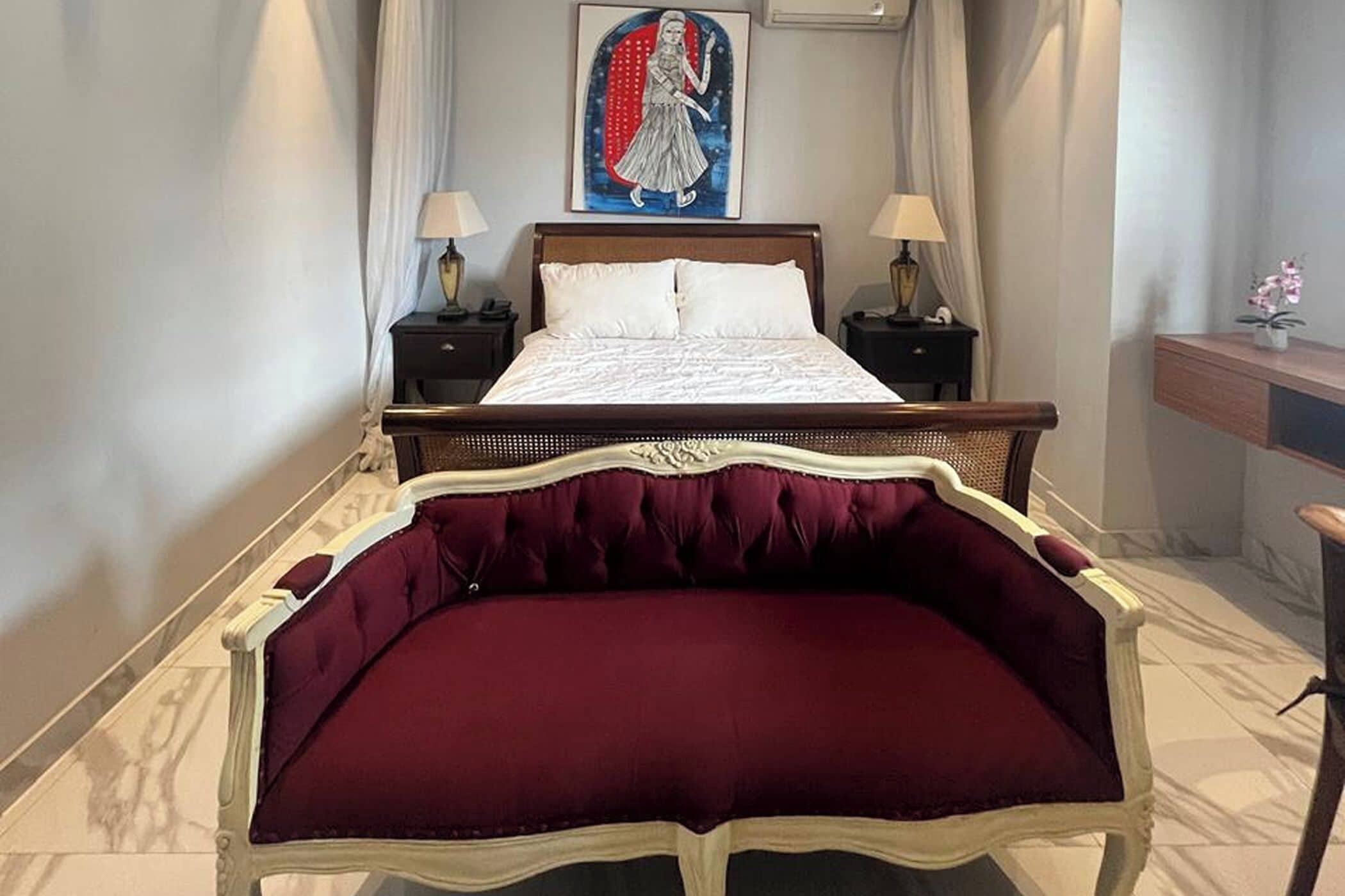 A hotel room with a large bed, two bedside tables with lamps, a painting on the wall, and a desk with a plant.