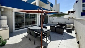 Een modern terras buiten, met een glazen deur die leidt naar een binnenruimte, een eettafel met stoelen en een grote parasol.