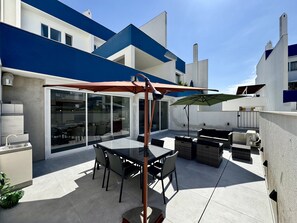 A modern outdoor patio with a glass door leading to an indoor area, a dining table with chairs, and a large umbrella. - VILAMOURA DELUXE 2 WITH POOL by HOMING (Vilamoura)