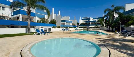A swimming pool with lounge chairs, palm trees, and modern buildings in the background.