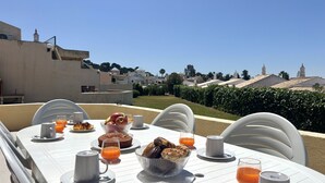 Mesa de jantar posta com itens de café da manhã, cadeiras de exterior e vista desobstruída para uma área residencial com casas e uma torre.