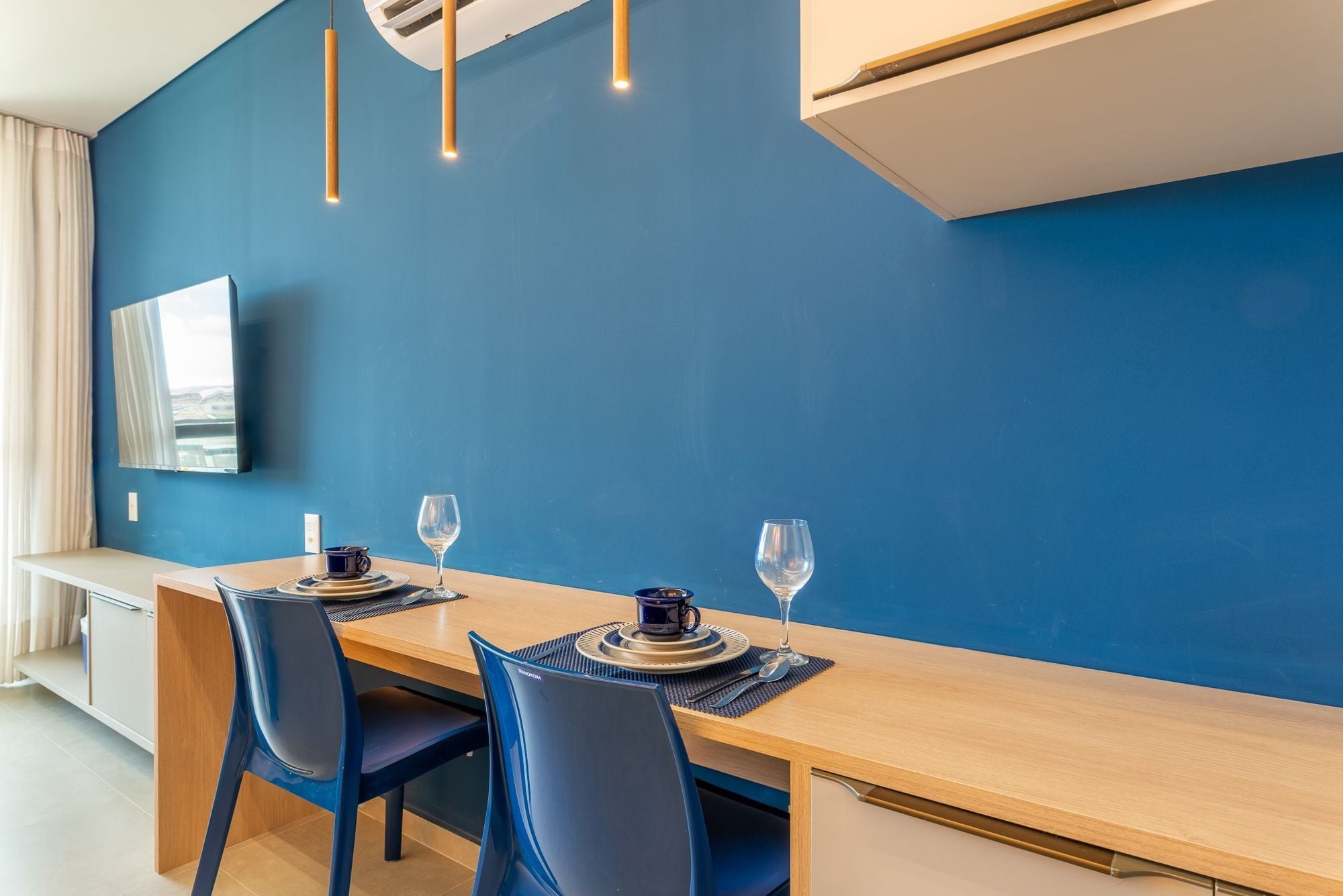 A modern dining area with a blue wall, a wooden table, two chairs, and a flat-screen TV mounted on the wall.