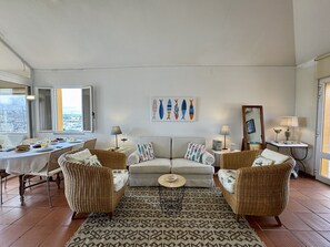 Ein Wohnzimmer mit einer Couch, zwei Sesseln, einem Esstisch mit Frühstück und Blick auf das Meer durch große Fenster.