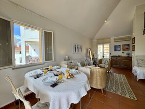 Ein Essbereich mit gedecktem Frühstückstisch, Korbstühlen, einem gemusterten Teppich und Blick ins Freie.