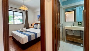 Habitación de hotel con una cama, un ventanal, un baño con lavamanos y espejo, y una puerta que da a otra habitación.