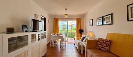 Una habitación con sofá, televisión, mesa con sillas, ventilador y vistas a través de una ventana.