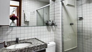 A bathroom with a granite countertop sink, a white toilet, a glass shower enclosure, and a mirror with a vase of flowers. - Cupe Beach Living Porto de Galinhas (Ipojuca)