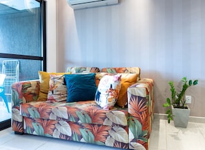 Un salón moderno con un sofá decorado con motivos de hojas tropicales, una planta en maceta y una ventana con una estantería metálica.